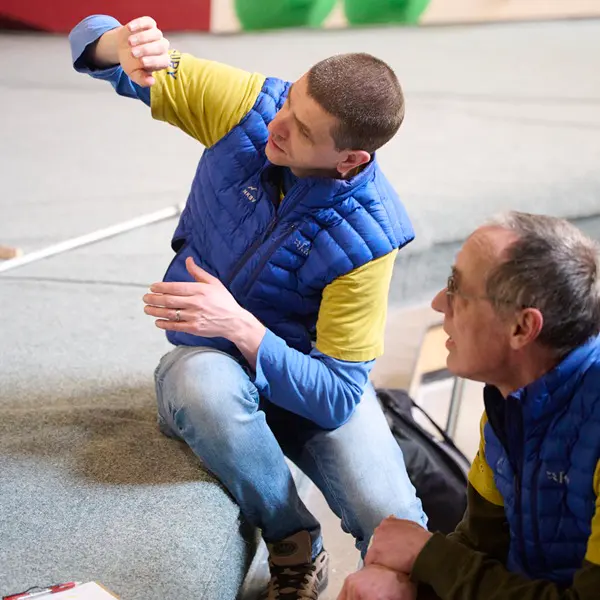 Twee mannen in blauwe vesten en gele shirts zitten binnen. De ene man wijst met zijn hand omhoog, terwijl de andere aandachtig toekijkt. Ze lijken in gesprek te zijn of iets te demonstreren.